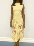 Woman wearing a light yellow sleeveless top and patterned skirt against a white wall.